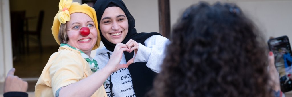En clown med röd näsa och en flicka i hijab formar ett hjärta tillsammans med varsin hand