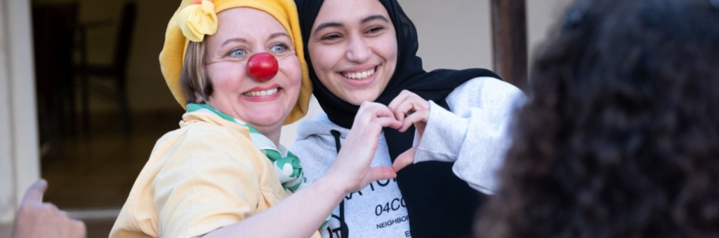 En clown i röd näsa och gul hatt står bredvid en tjej som ler och tillsammans formar dom sina händer till ett hjärta