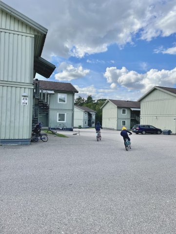 två barn cyklar i hög fart på en asfaltsplan mellan träbyggnader
