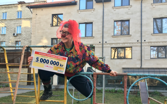 En clown med röd näsa ler mot kameran och håller i en check med summan 5 miljoner kronor