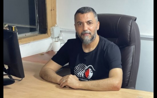 Mustafa Sheta sitter vid ett skrivbord och tittar in i kameran. Han har kortklippt hår, grått skägg och en svart t-shirt