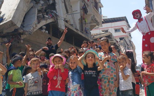 En grupp barn står tillsammans med en artist på styltor och gör tummen upp. I bakgrunden sönderbombade hus